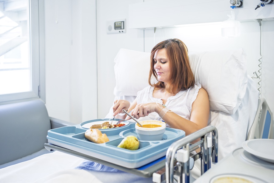 Paciente en cama de hospital comiendo menú balanceado con sopa, pan, fruta y proteínas, siguiendo alimentación postoperatorio cirugía estética recomendada por especialistas.