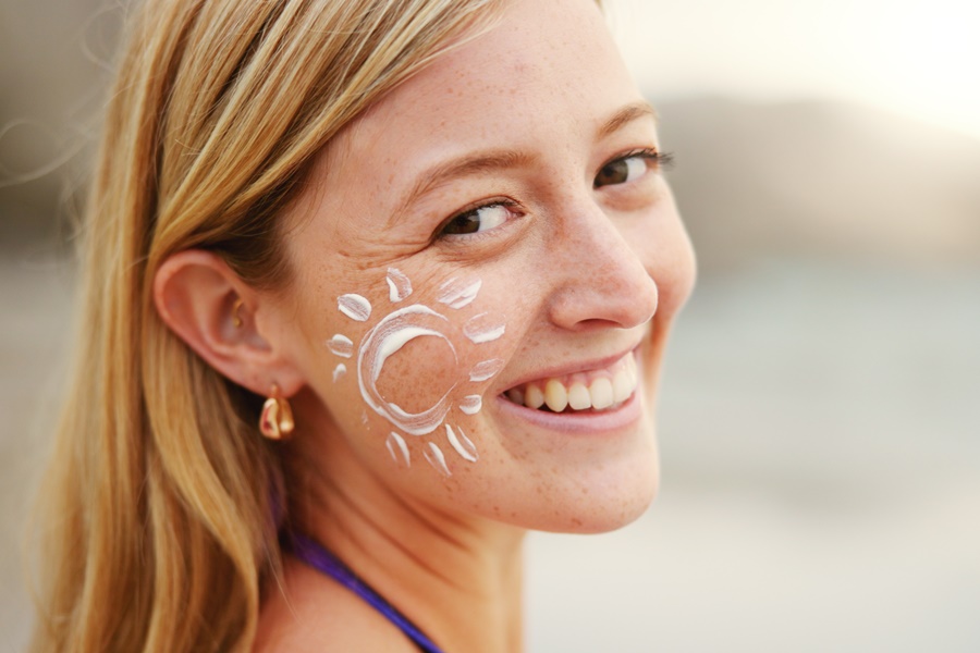 Mujer joven con protector solar dibujado como un sol en la mejilla, sonriendo al aire libre. Imagen ideal para ilustrar la importancia de usar fotoprotección para prevenir el envejecimiento prematuro de la piel.