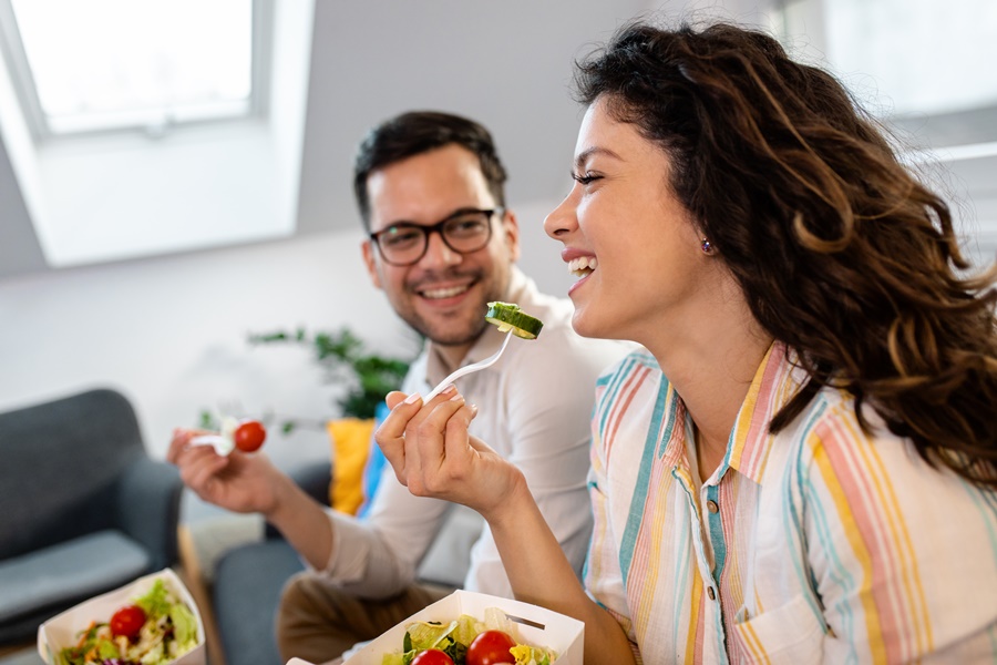 Mujer sonriendo mientras come una ensalada, acompañada de un hombre. Representa una alimentación equilibrada como parte fundamental para prevenir el envejecimiento prematuro de la piel.