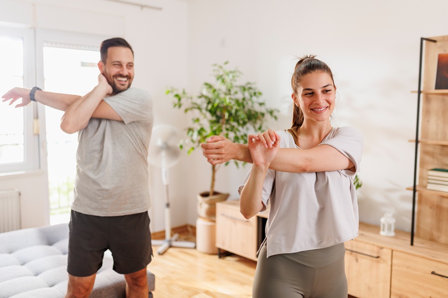 Pareja joven realizando estiramientos en casa, en un ambiente relajado. Imagen relacionada con la actividad física regular como método para prevenir el envejecimiento prematuro de la piel.