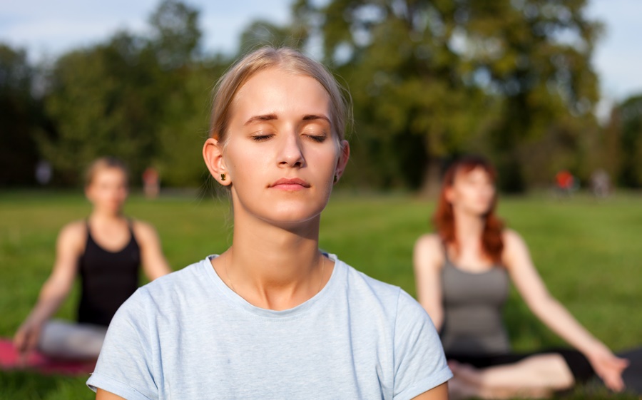Mujer practicando meditación al aire libre con los ojos cerrados, en grupo. Representa el manejo del estrés como hábito para prevenir el envejecimiento prematuro de la piel.