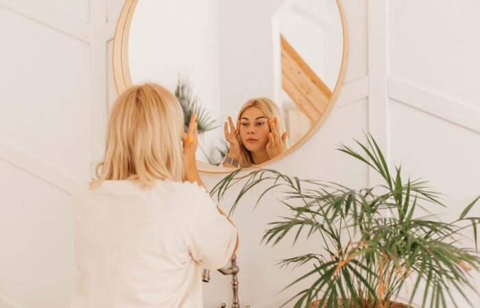 Mujer rubia frente al espejo aplicando crema en el rostro, representando la reflexión personal previa a una cirugía plástica y la gestión de expectativas realistas.