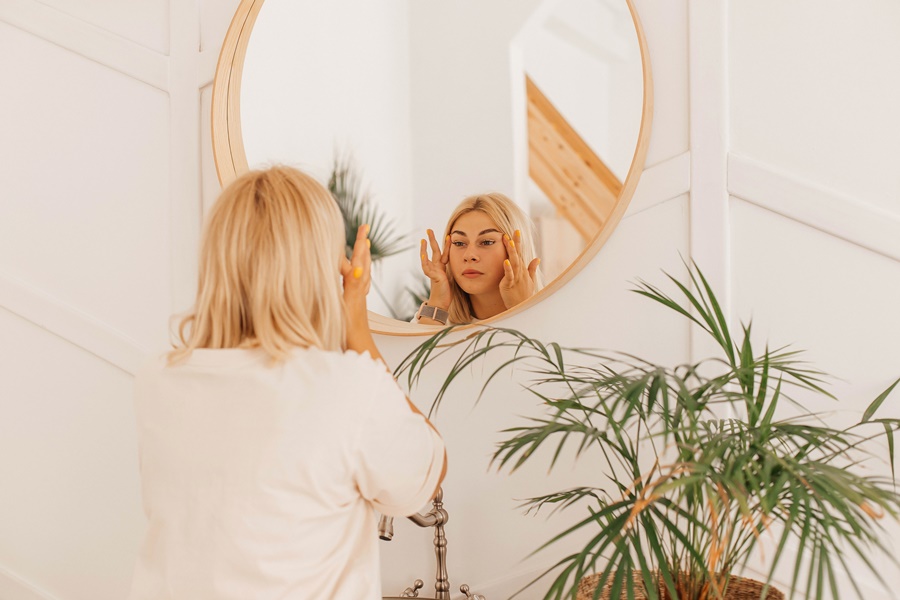 Mujer rubia frente al espejo aplicando crema en el rostro, representando la reflexión personal previa a una cirugía plástica y la gestión de expectativas realistas.