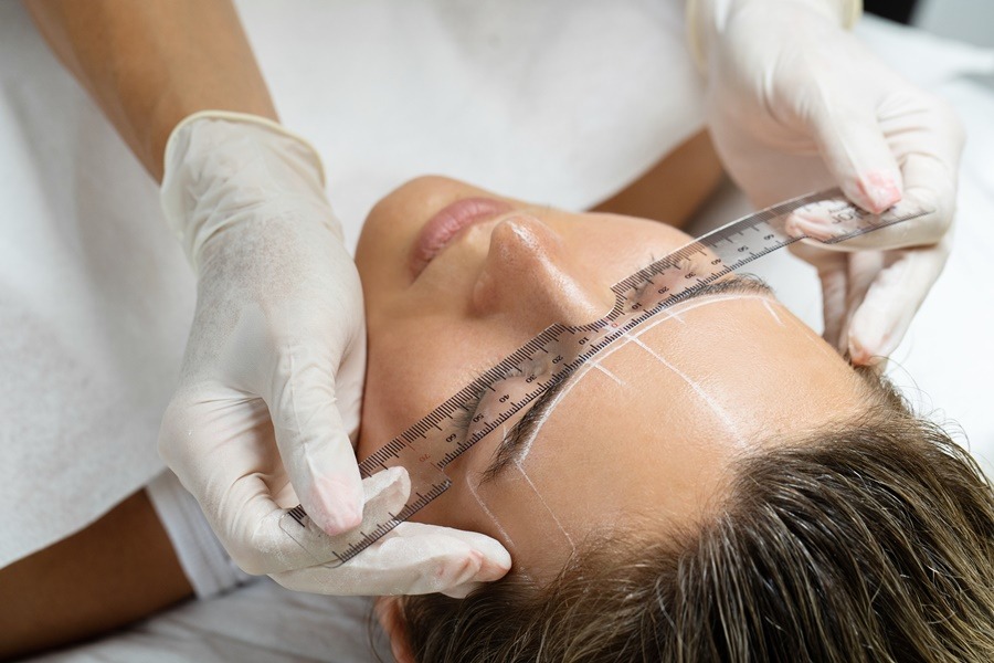 Medición de la línea de las cejas en un rostro femenino antes de un lifting directo de cola de cejas, con guantes y regla transparente sobre la frente.