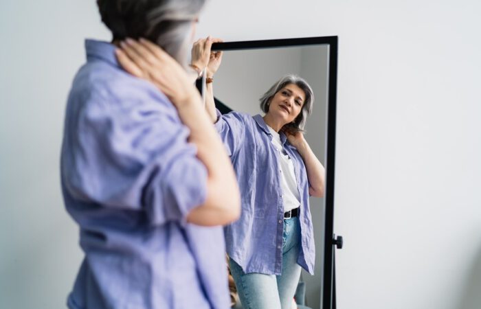 Mujer en etapa de menopausia observándose frente al espejo con actitud confiada, vestida con ropa casual y fondo neutro, imagen representativa del vínculo entre menopausia y cirugía estética.