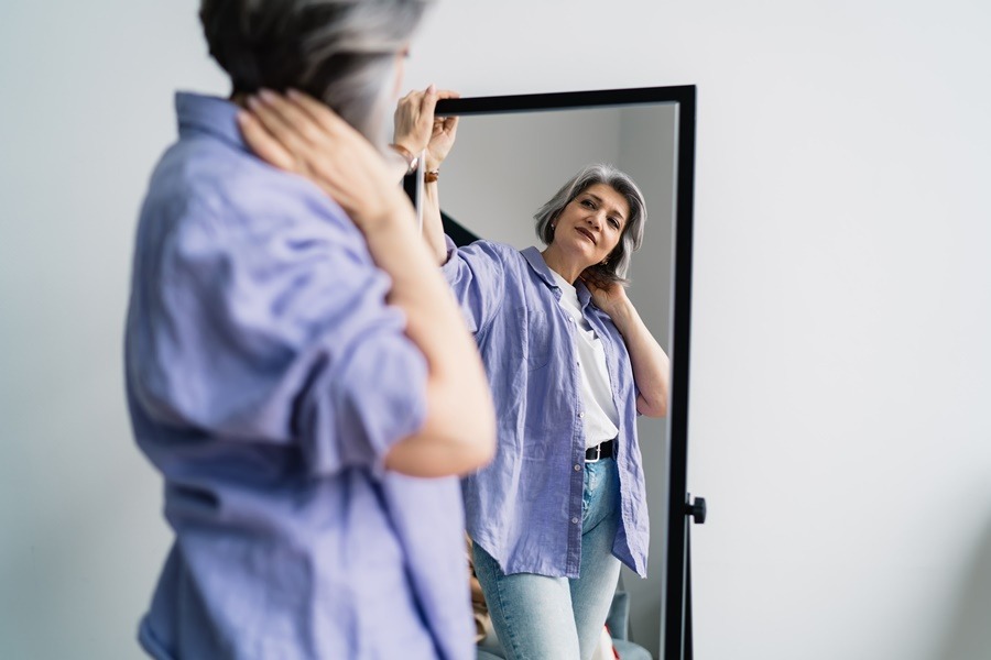 Mujer en etapa de menopausia observándose frente al espejo con actitud confiada, vestida con ropa casual y fondo neutro, imagen representativa del vínculo entre menopausia y cirugía estética.